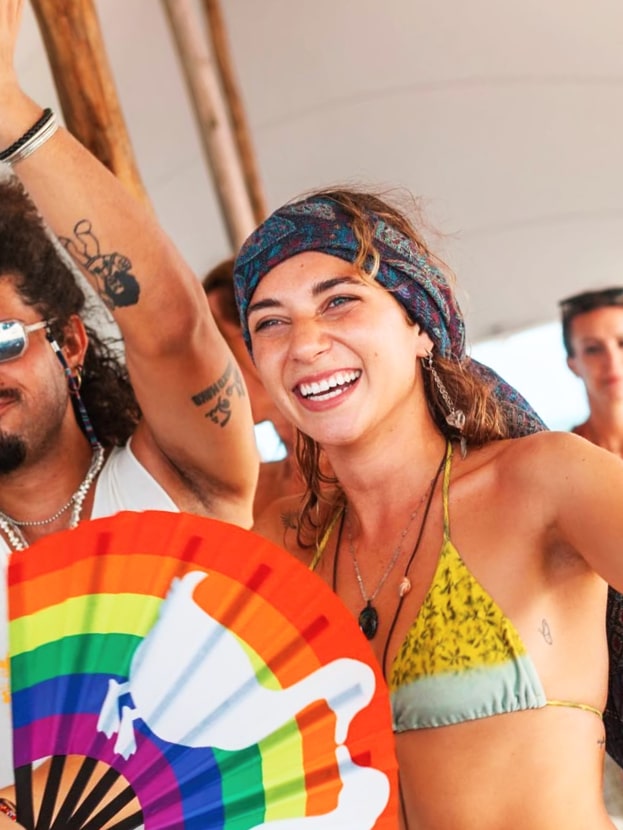 Smiling woman in festival outfit holding rainbow fan surrounded by friends—vibrant event and lifestyle