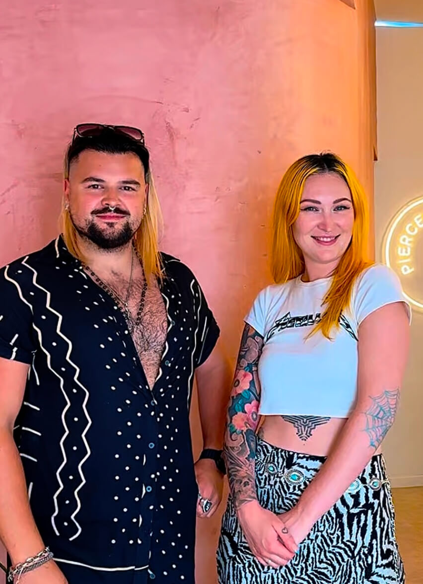 Smiling man and woman with tattoos posing stylishly against a pink wall and neon sign—modern lifestyle and brand content in Ibiza by vIBZ Marketing