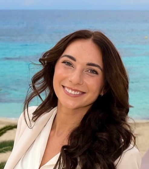 Young woman with long dark hair in a white blazer, smiling in front of the sea in Ibiza. Member of the vIBZ Marketing team.