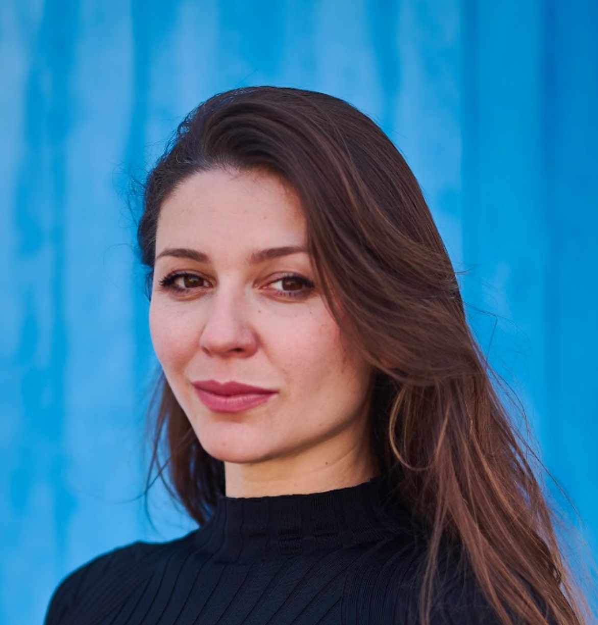 Portrait of a woman with long brown hair in a black top, part of the vIBZ Marketing creative team, standing in front of a bright blue background.