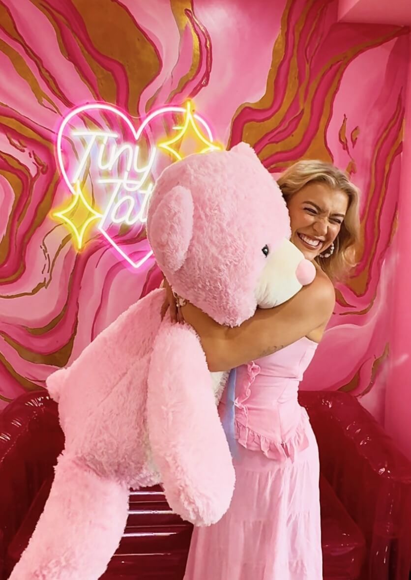 “Smiling woman hugging a giant pink teddy bear in front of neon sign ‘Tiny Tattoo’—playful lifestyle and branding content creation in Ibiza