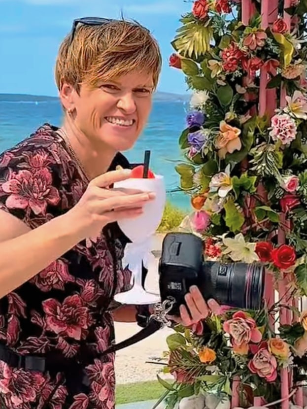 Smiling woman with camera and cocktail at a floral-decorated beach wedding in Ibiza—professional event photography by vIBZ Marketing.