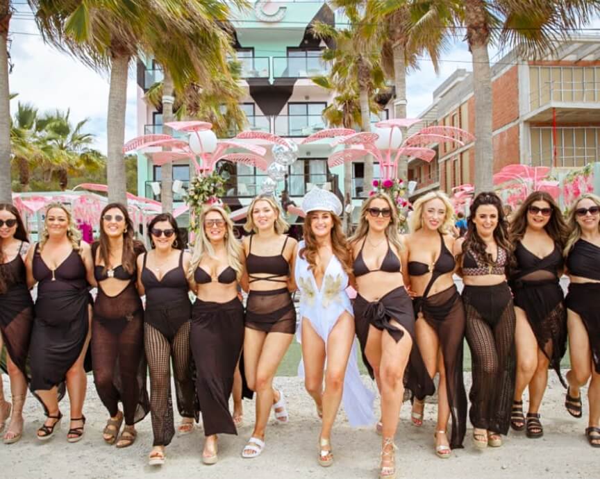 Group of women in stylish black outfits and one in a white embellished bodysuit celebrating together at a glamorous Ibiza pool party—event and lifestyle content by vIBZ Marketing.