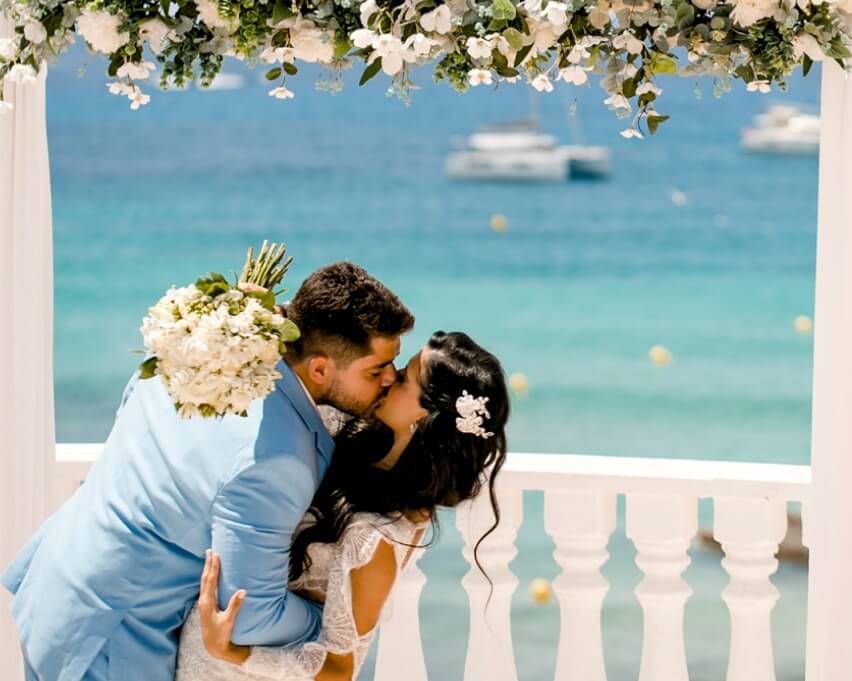 Romantic Ibiza beach wedding – newlyweds kiss under floral arch with Mediterranean sea view.