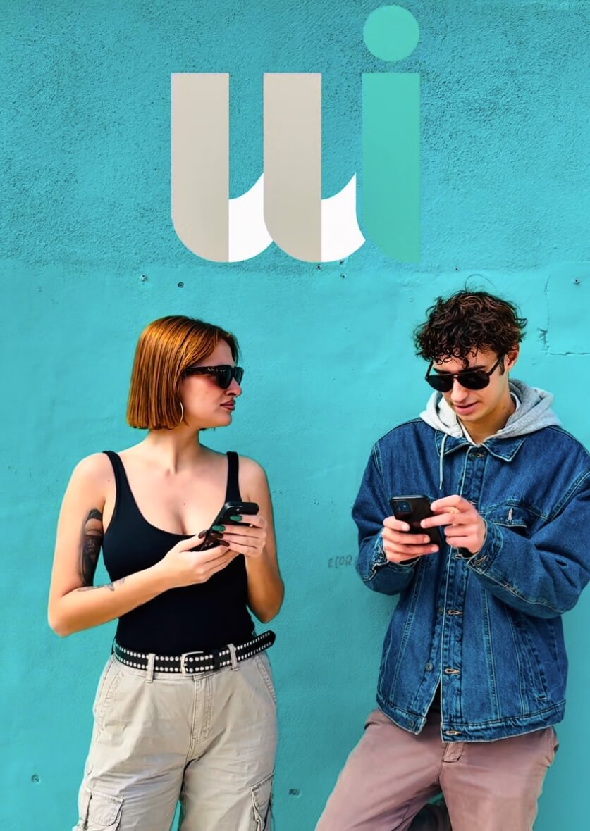 Young man and woman in casual outfits holding smartphones in front of turquoise wall with WI logo—urban lifestyle and brand content creation in Ibiza.
