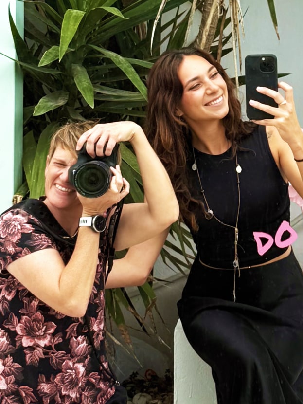 Two women laughing while capturing photos with professional camera and smartphone—creative behind-the-scenes social media content in Ibiza